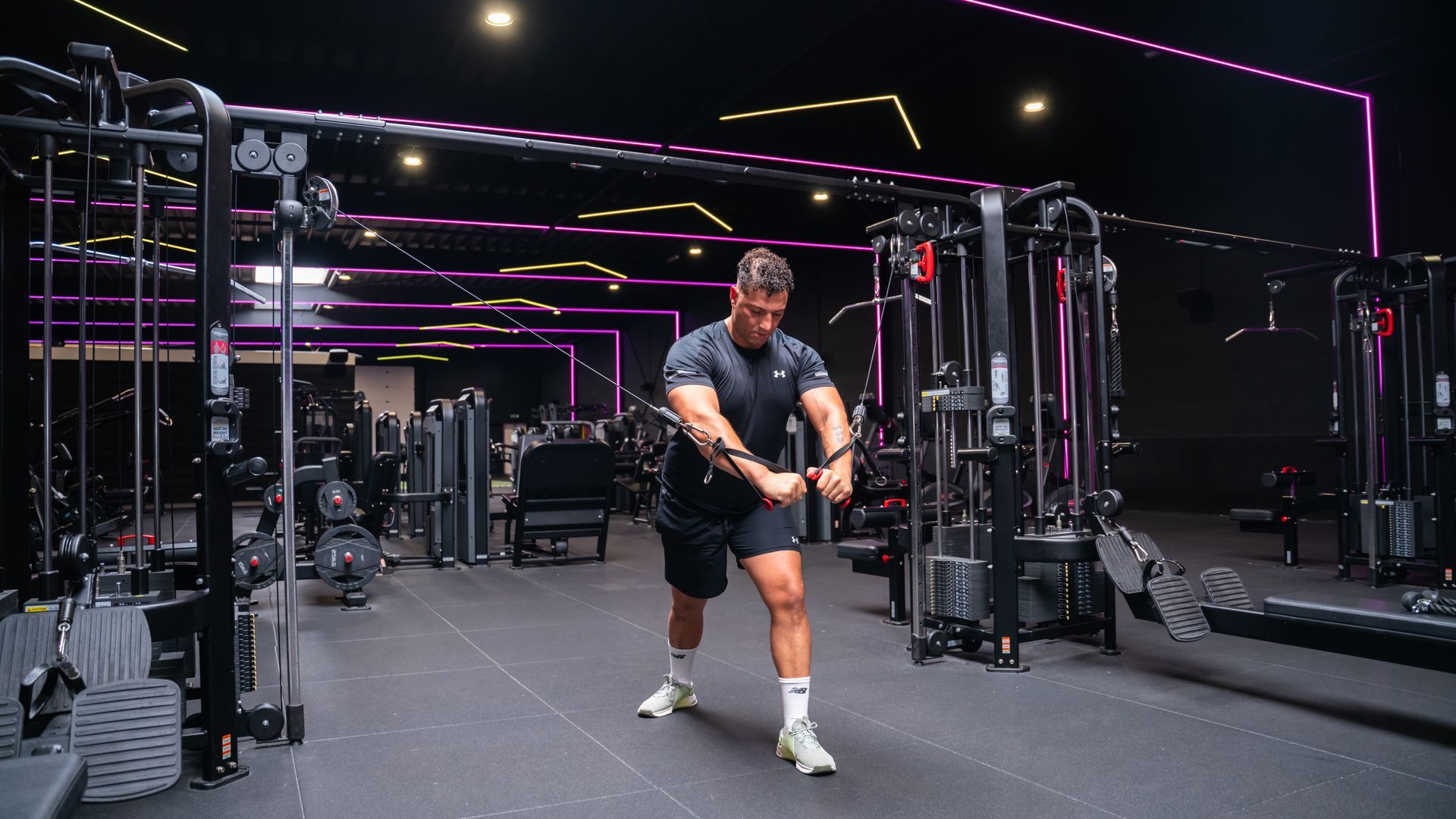 Een man is aan het trainen met een kabelmachine in een sportschool met neonverlichting, gekleed in zwarte sportkleding.