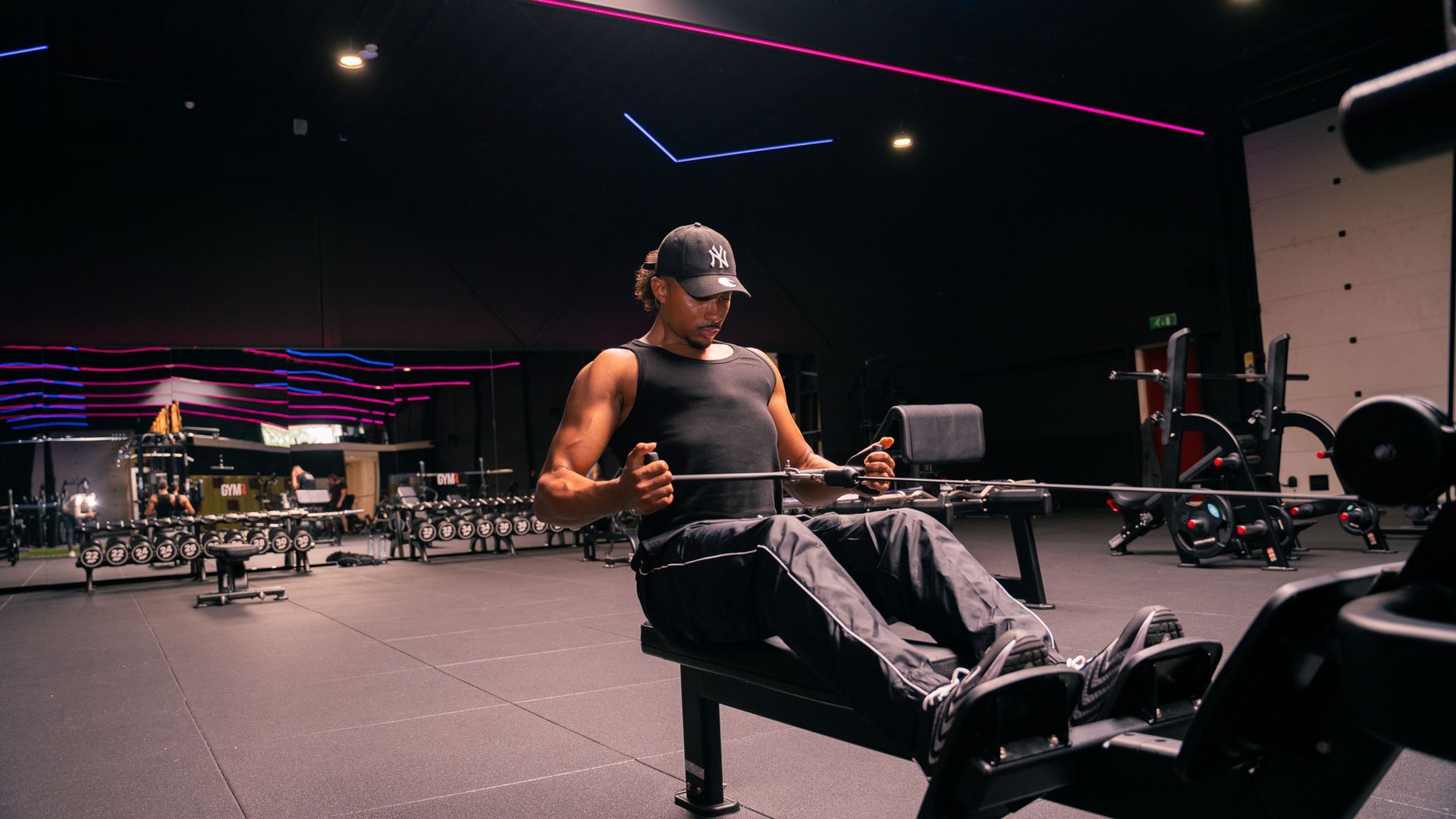 Man using a chest press machine in a gym, wearing a yellow shirt against a blue-lit brick wall.