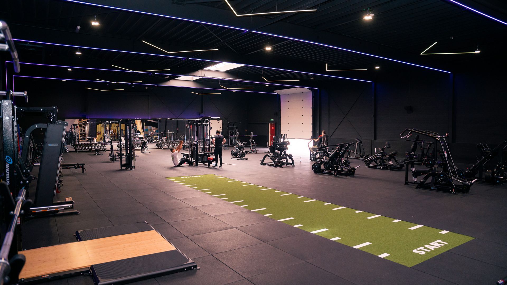 Empty modern gym with weight machines, black floors, and a green turf strip under dim lighting