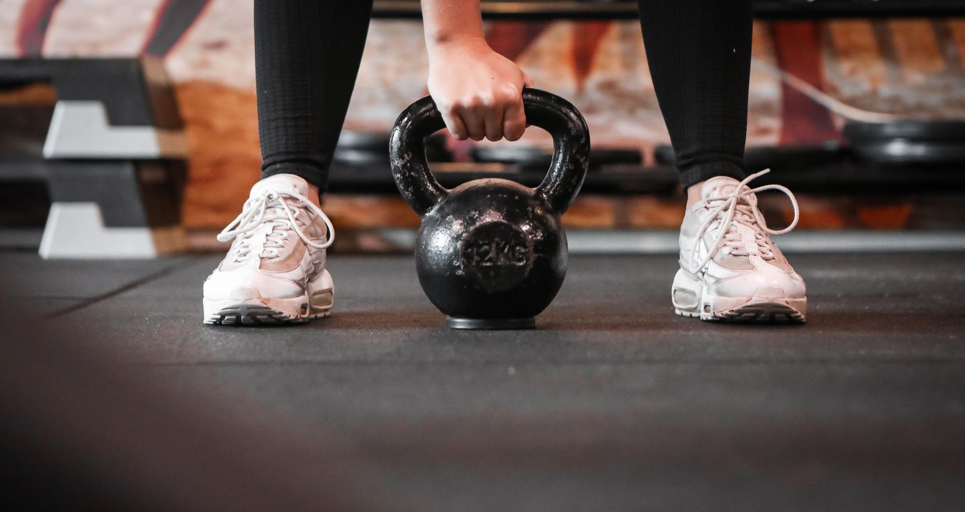 Person lifting a kettlebell in a gym, wearing white sneakers on a rubber floor.