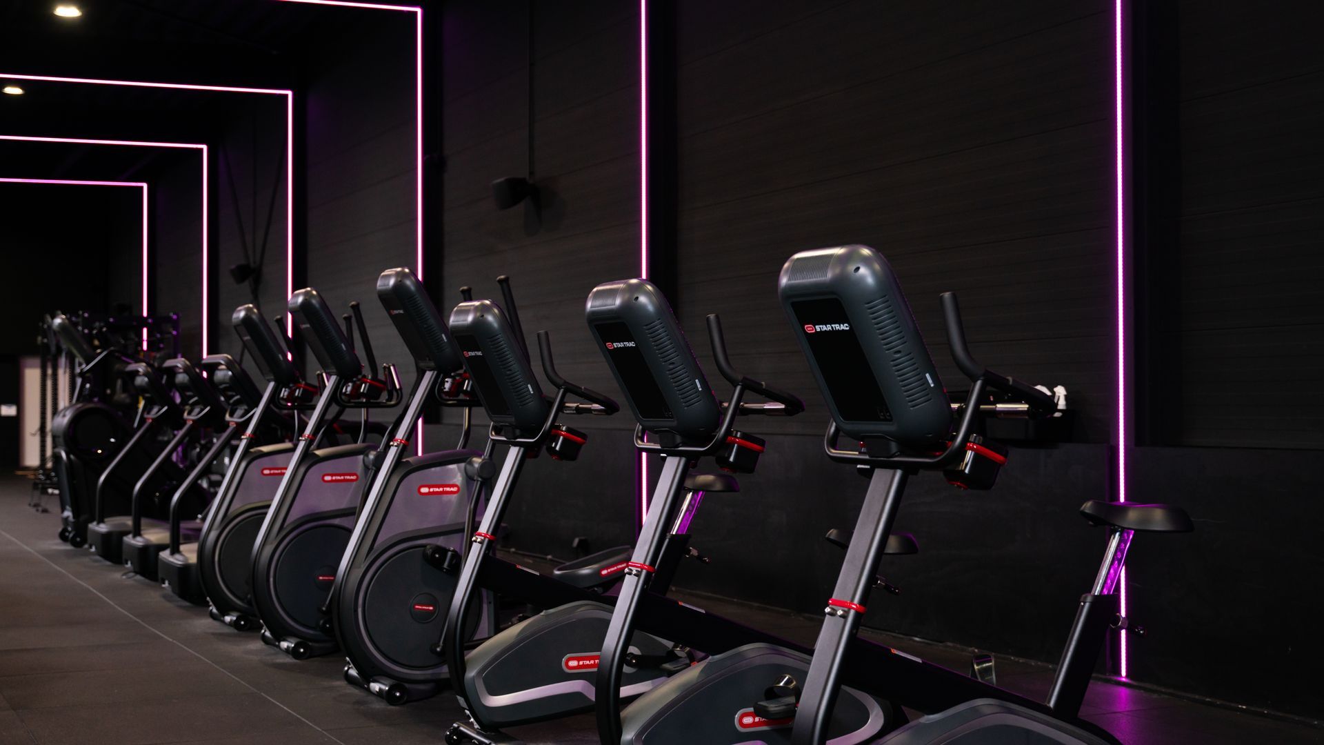Row of black stationary exercise bikes in a dim gym with pink neon lighting