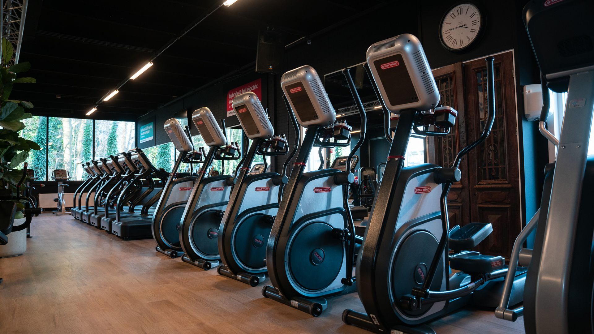 Row of black elliptical machines in a gym with wood floors, mirrors, and wall clocks