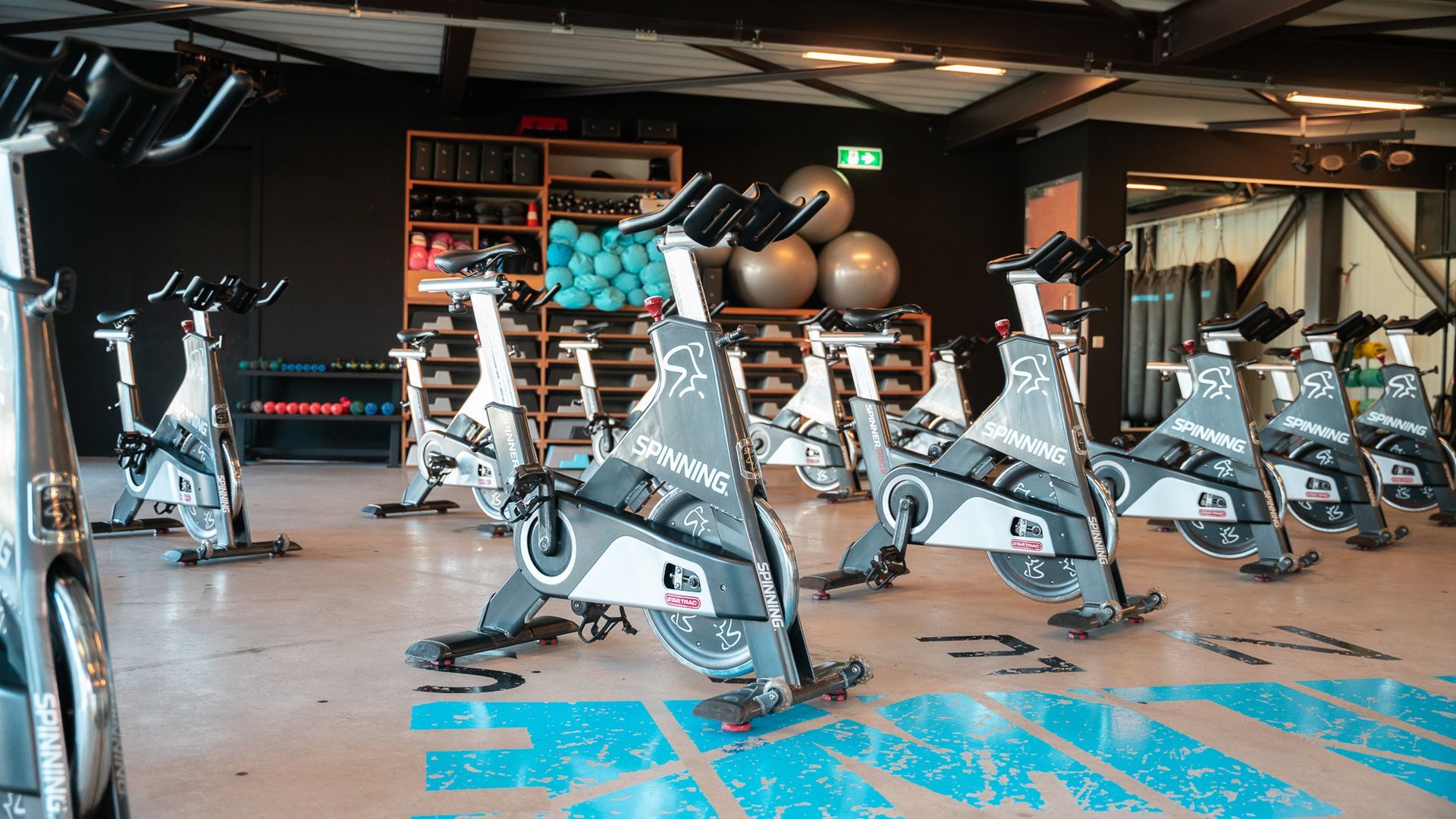 Row of stationary exercise bikes in a modern gym with dumbbells and workout balls in the background