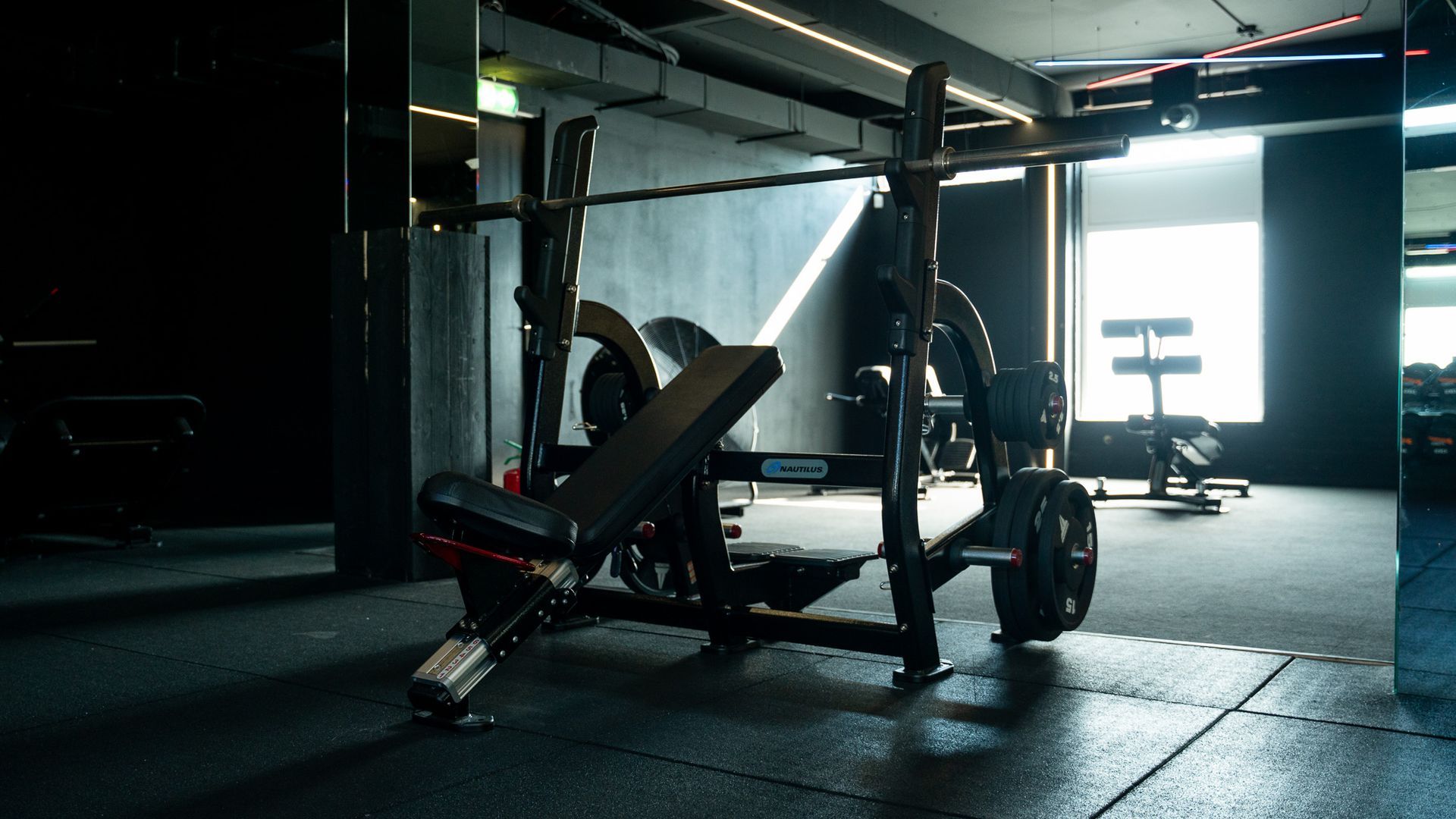 Gym bench press station in a dimly lit fitness room with equipment and sunlight from a window