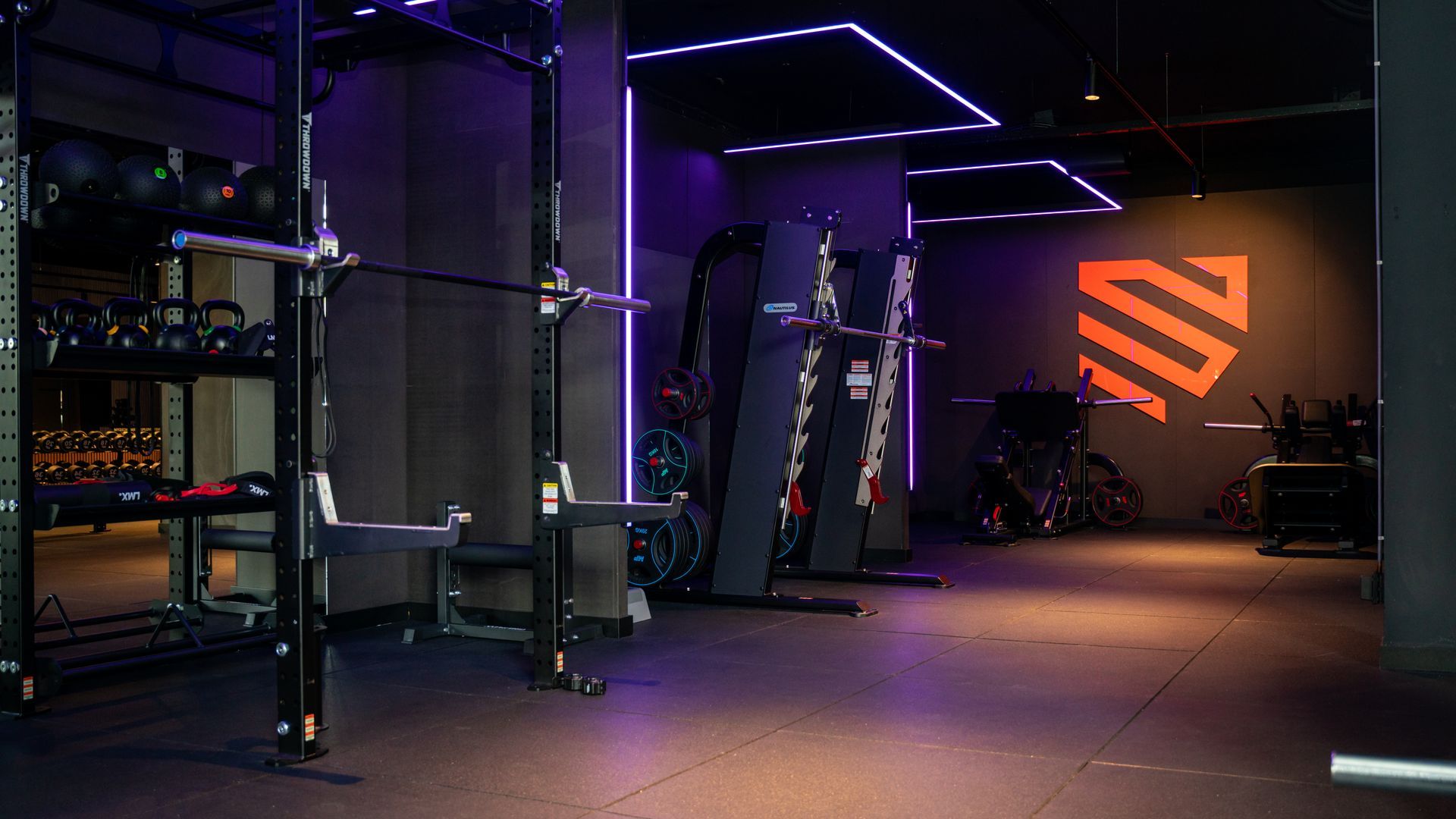 Modern gym interior with purple LED lighting, weight equipment, and an orange logo on the back wall