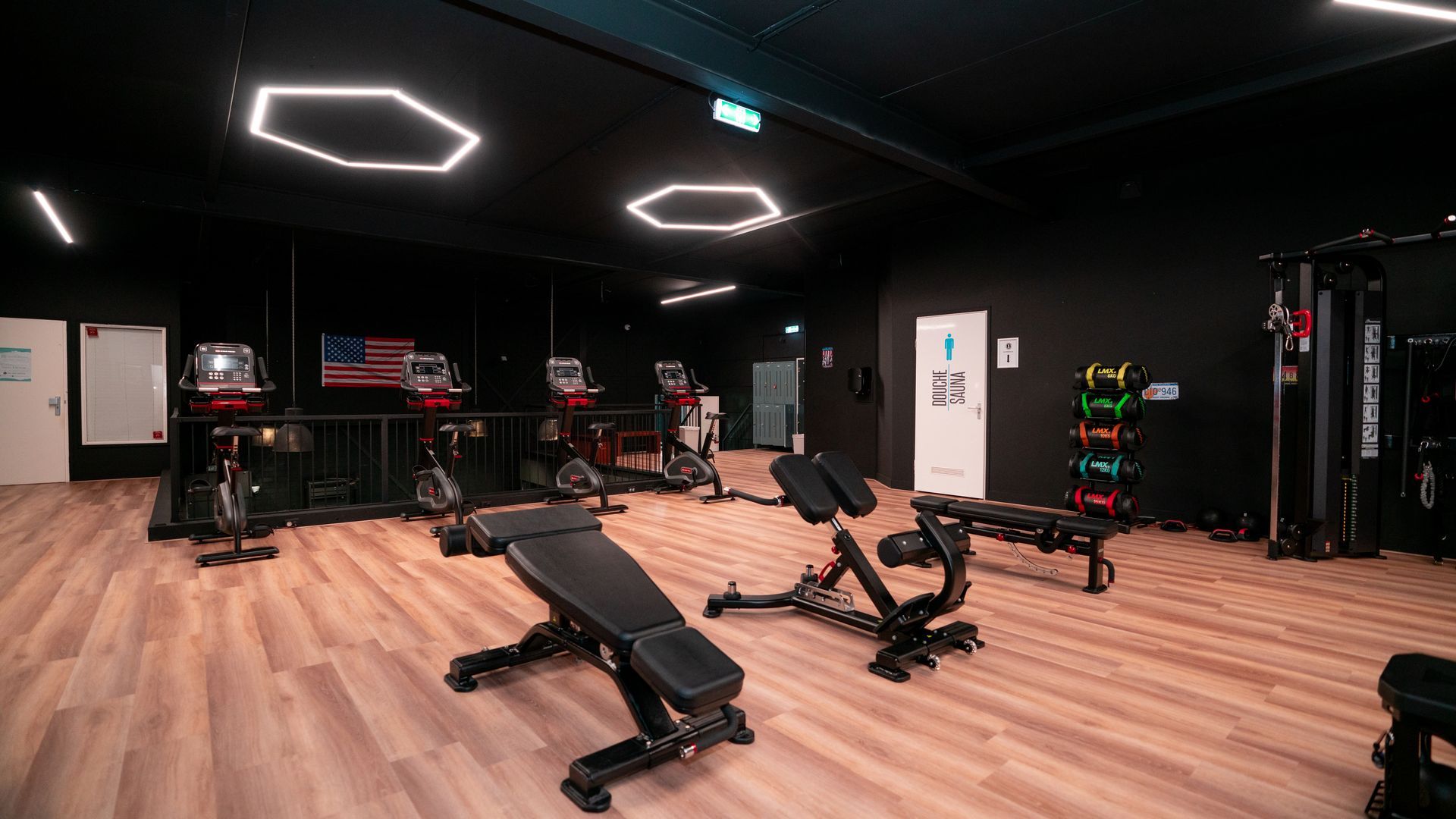 Modern gym interior with black walls, wood floors, benches, cardio machines, and geometric ceiling lights