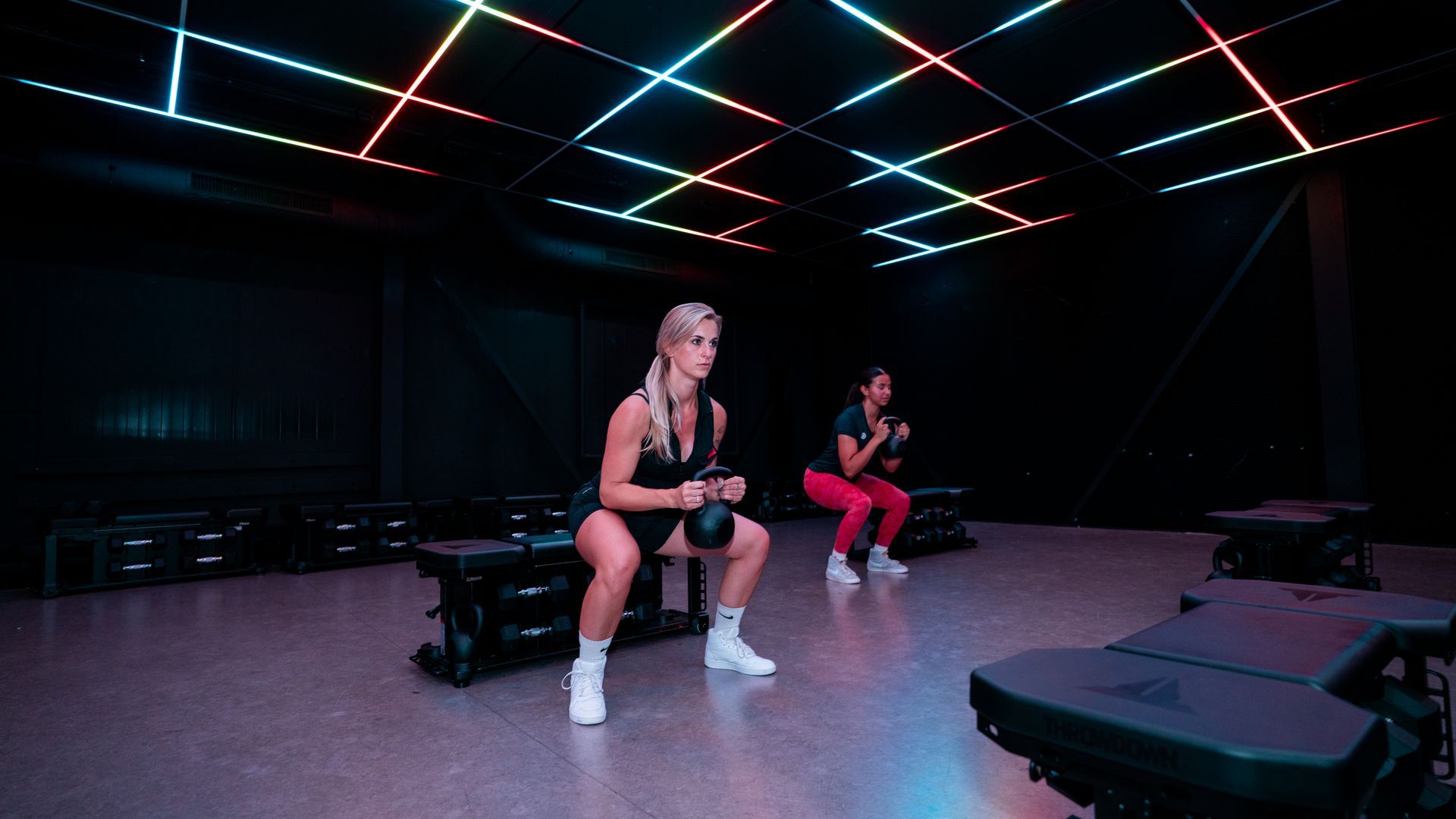 Two people sit on benches in a neon-lit gym or studio, with colorful ceiling lights overhead.