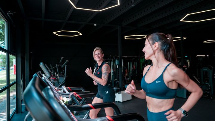 Two women in teal workout clothes on treadmills in a dark gym, smiling and gesturing during a workout.