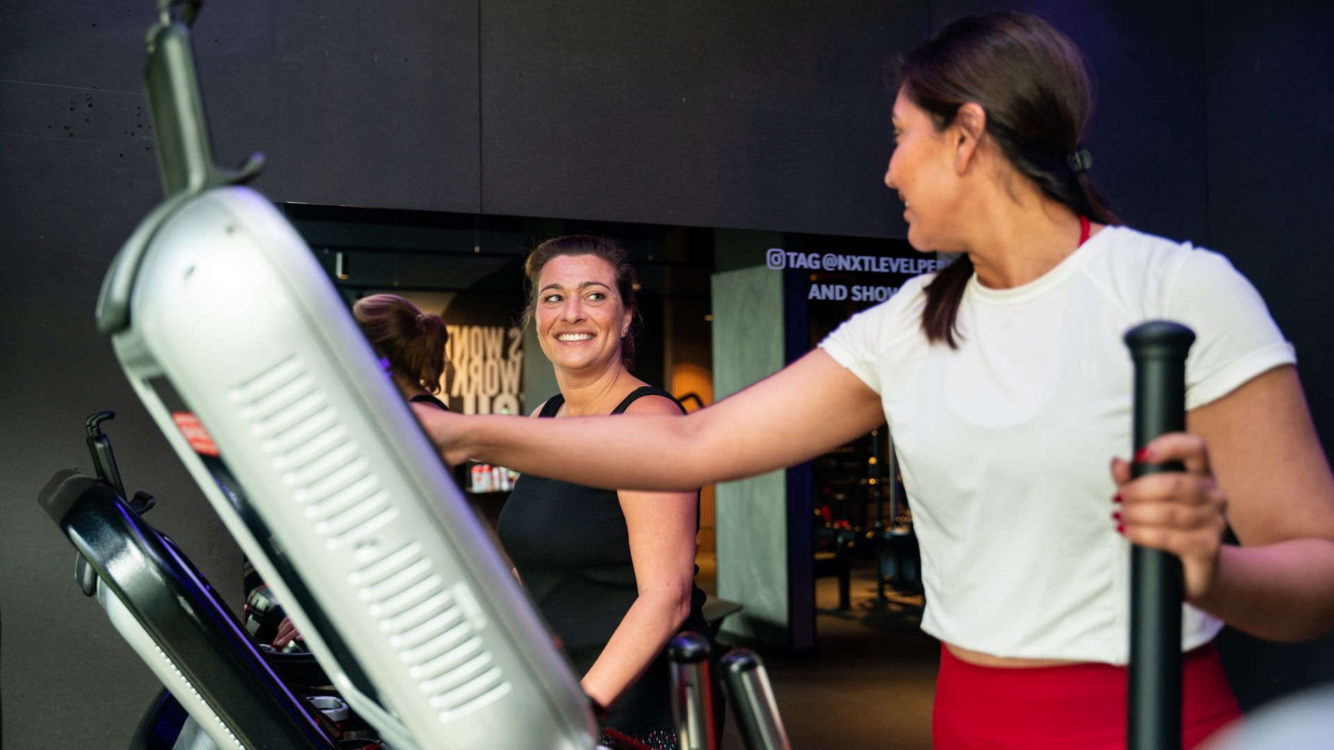 Two women using cardio machines in a gym, one smiling and reaching toward the console.