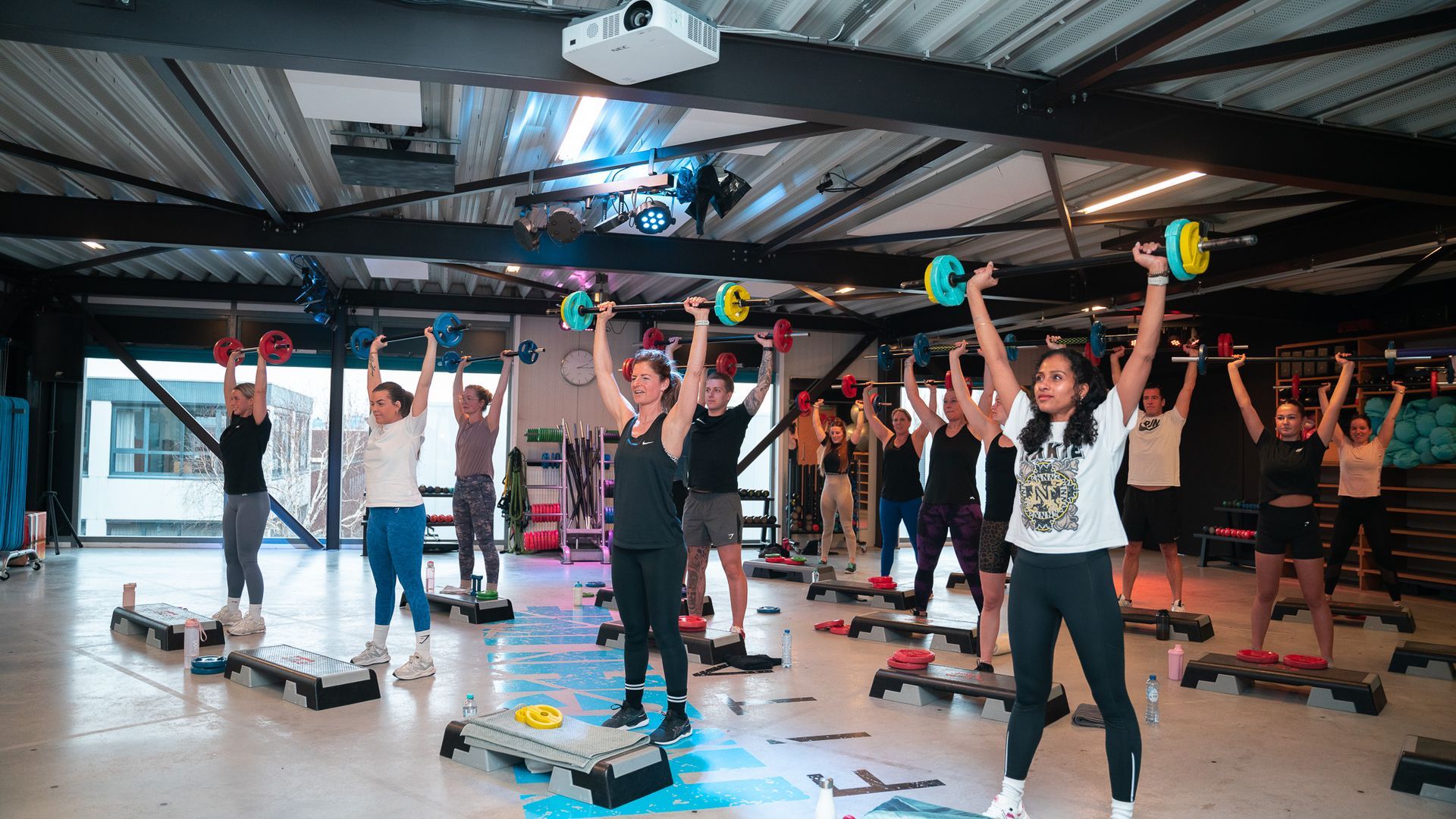 Fitness class participants in a gym lift colorful dumbbells overhead in unison.