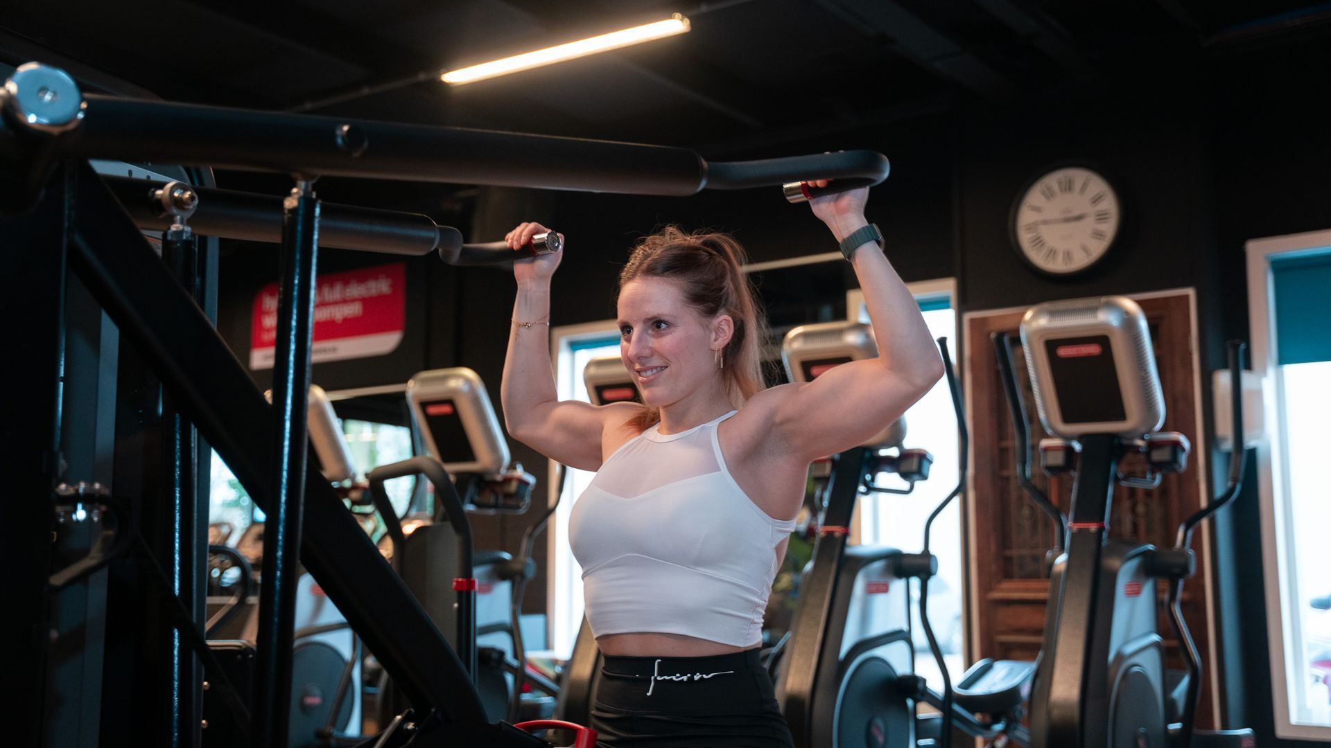 Woman lifting weights in a gym, standing between cardio machines with arms raised and focused expression