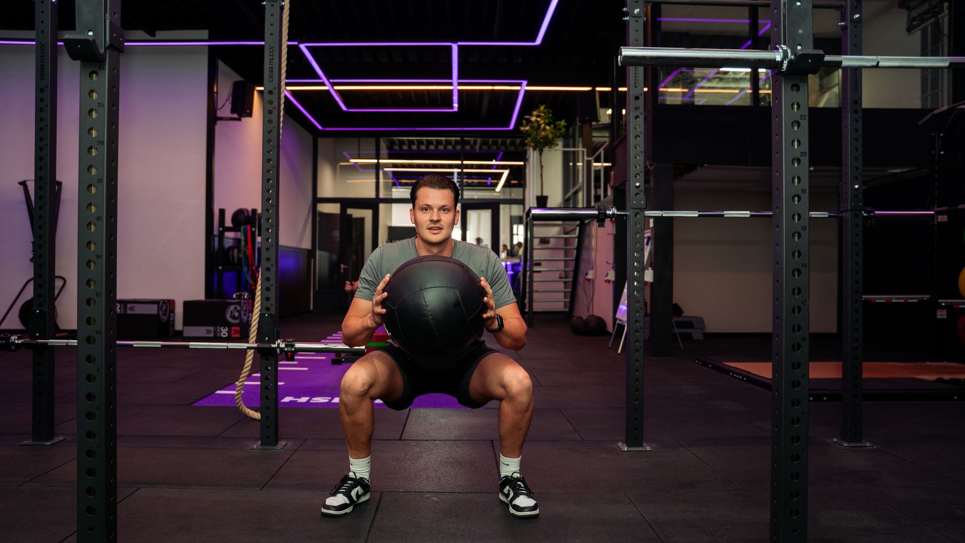 Man holding a medicine ball in a gym, seated between weight racks under purple neon lights