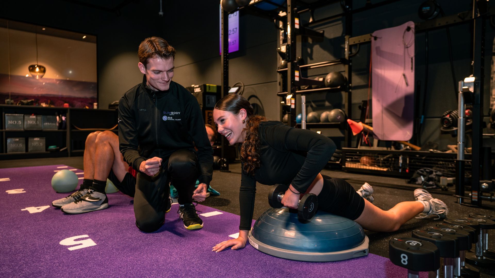 Two people exercising on balance trainers in a gym with purple carpet and equipment in the background