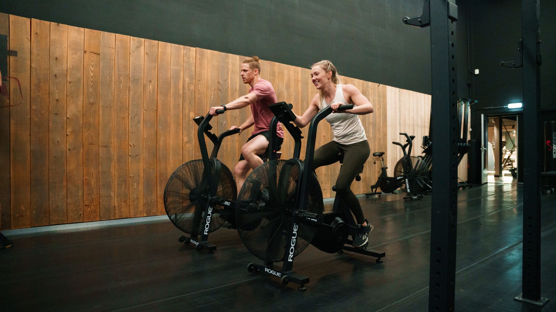 Two people riding stationary bikes in a dim indoor studio, one in pink and one in tan.