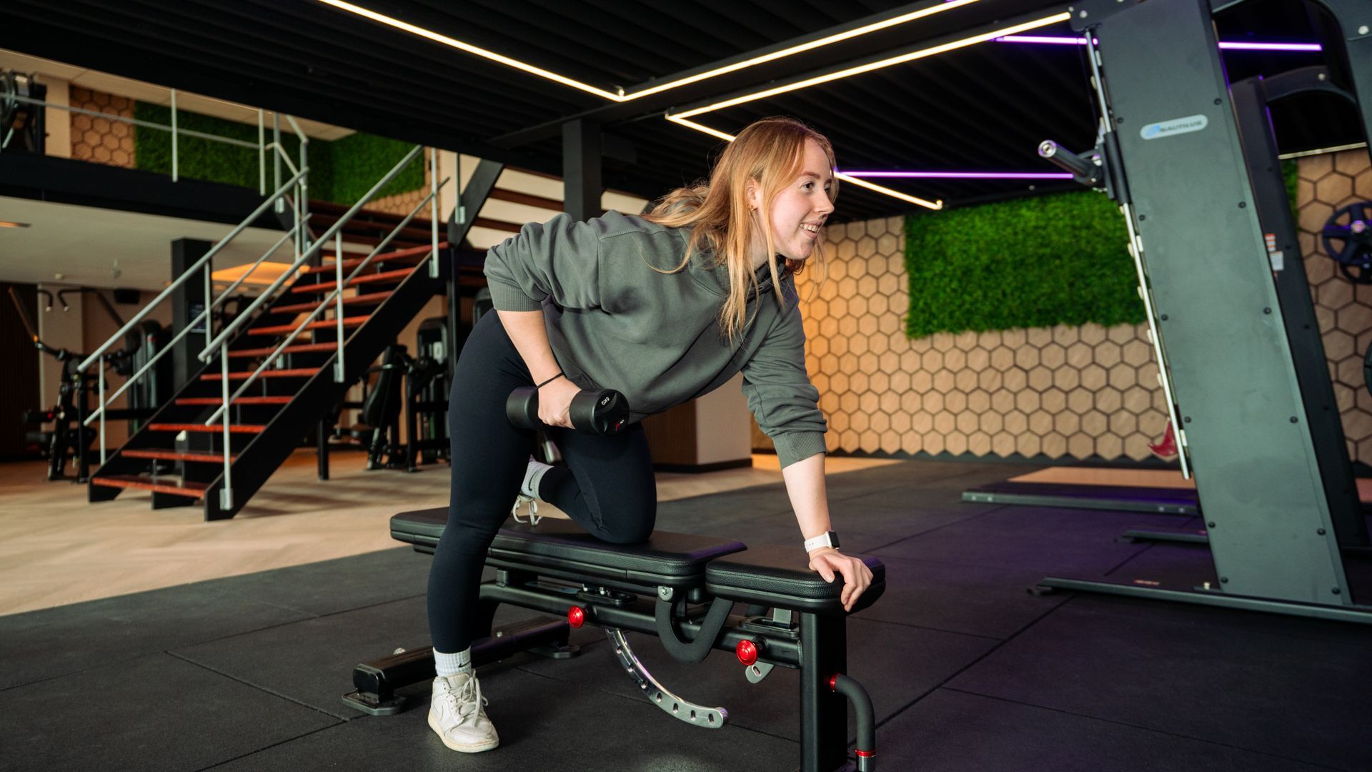 Woman adjusting a dumbbell on a bench in a modern gym, with stairs and hexagonal wall panels behind her