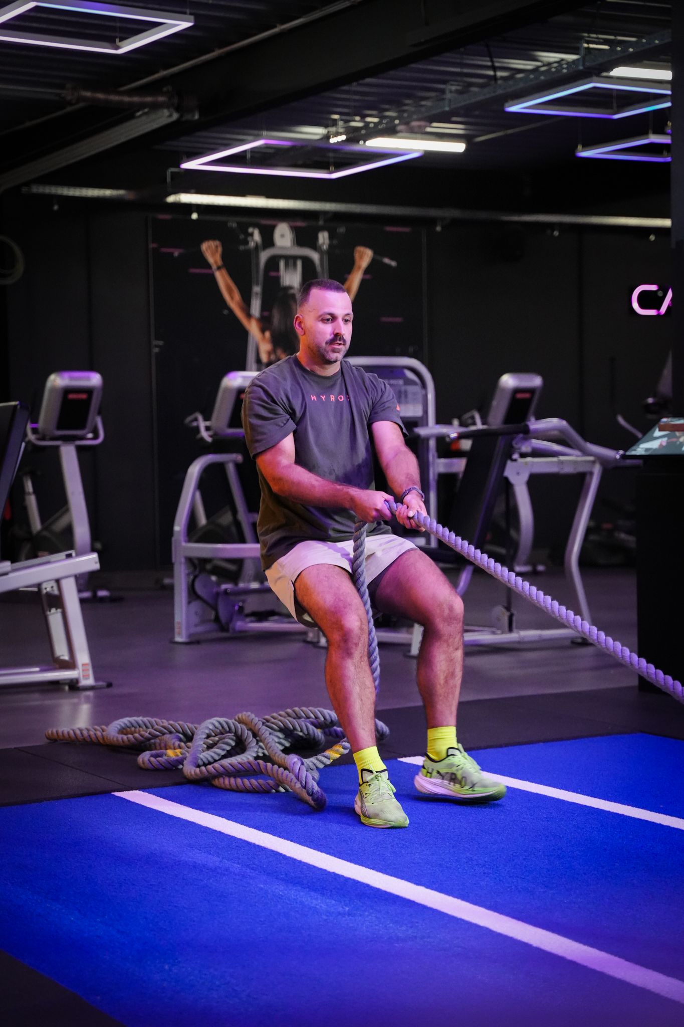 Person doing battle rope exercise in a gym, seated on a blue turf track near treadmills.
