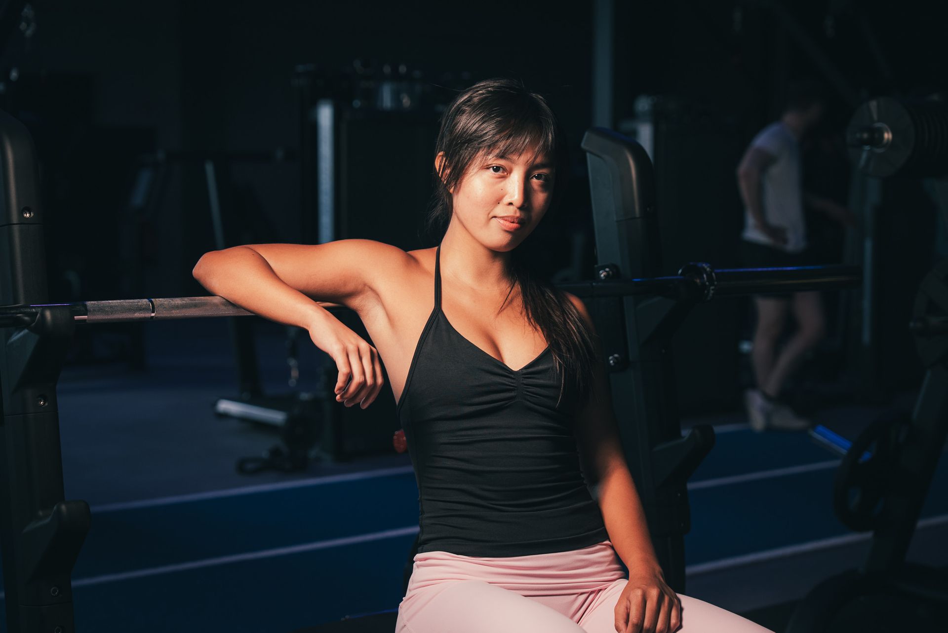 Woman in a black tank top sits on a bench in a dim gym, leaning on a barbell rack.