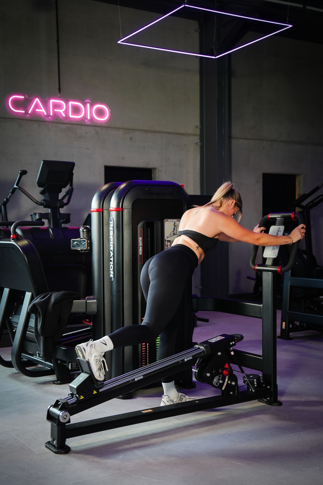 Person using a gym machine in a dark fitness center with neon pink “CARDIO” sign.
