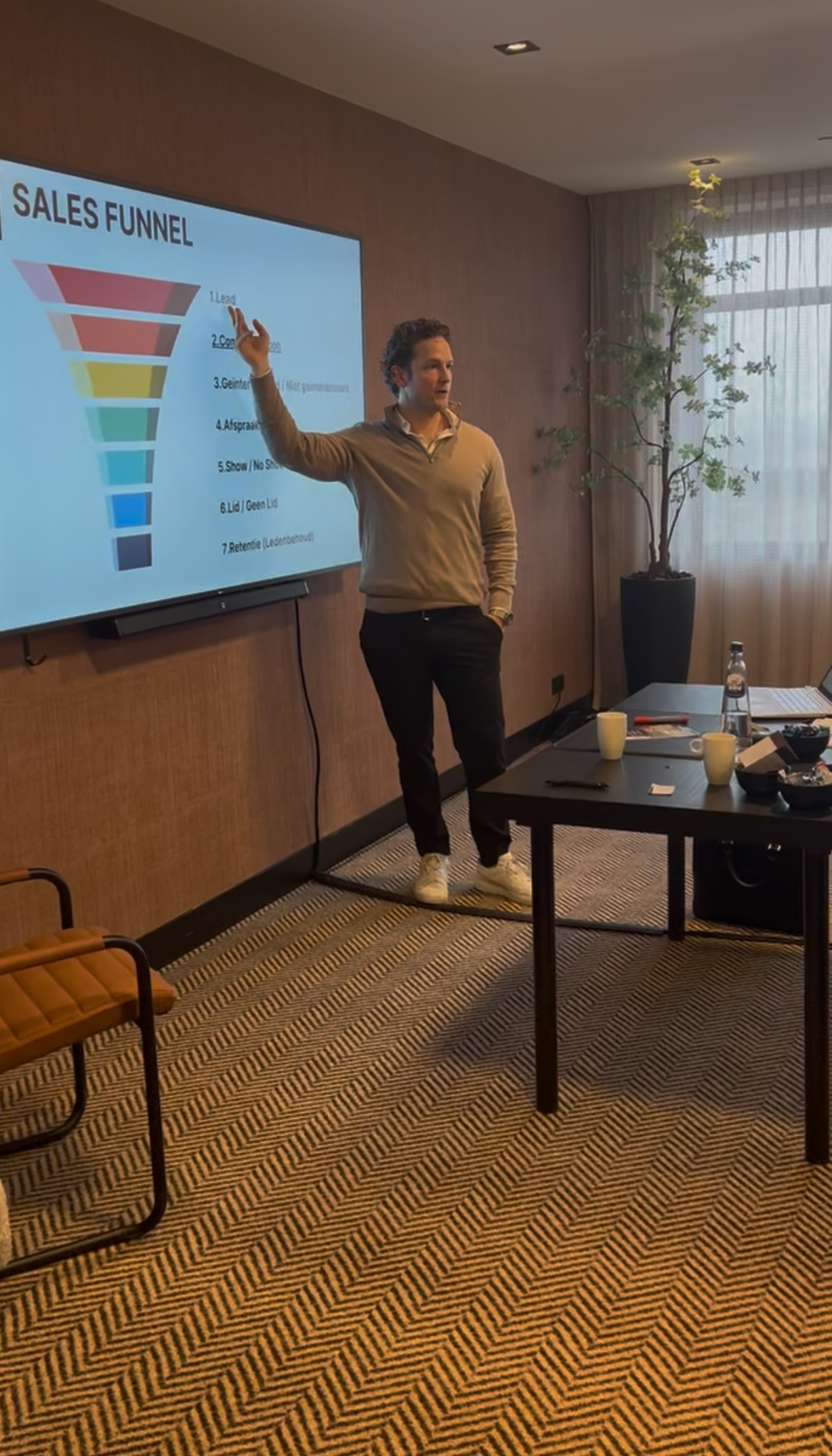Presenter beside sales funnel slide in a meeting room, pointing at a screen during a presentation.