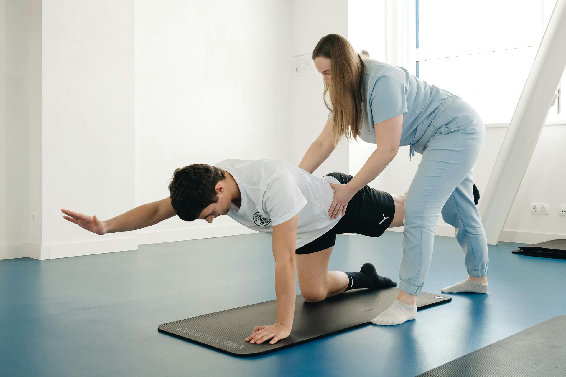 Physical therapy session: one person assists another doing a balance exercise on a mat in a bright studio