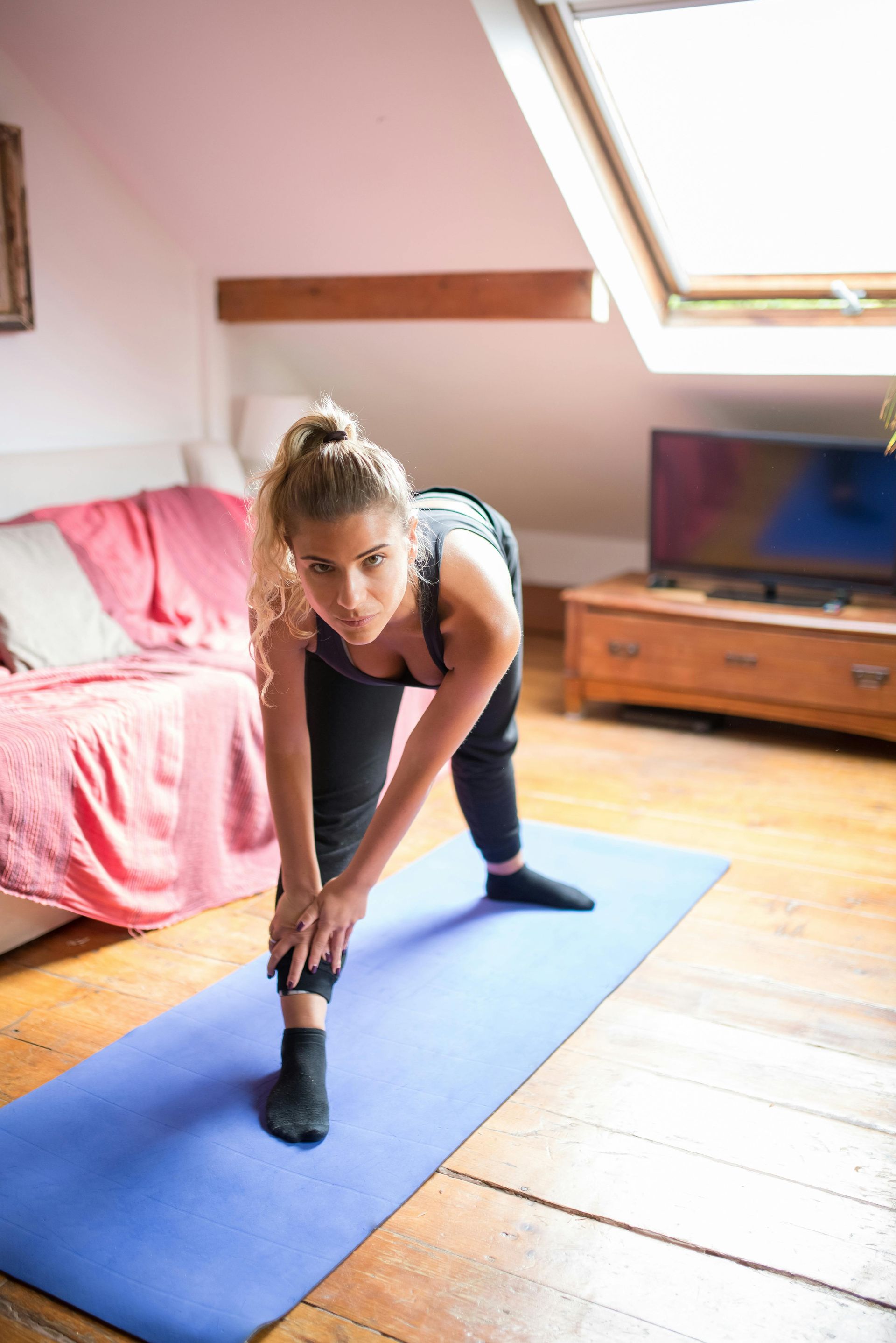 Woman stretching on a blue yoga mat in a sunlit attic room.