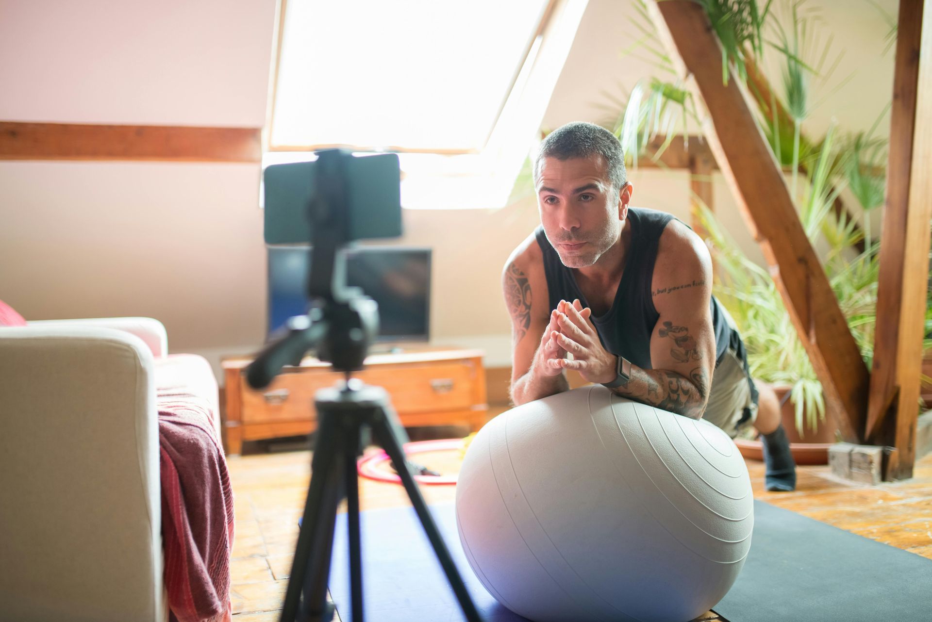 Person leaning on a yoga ball, filming a workout on a tripod in a bright attic room.