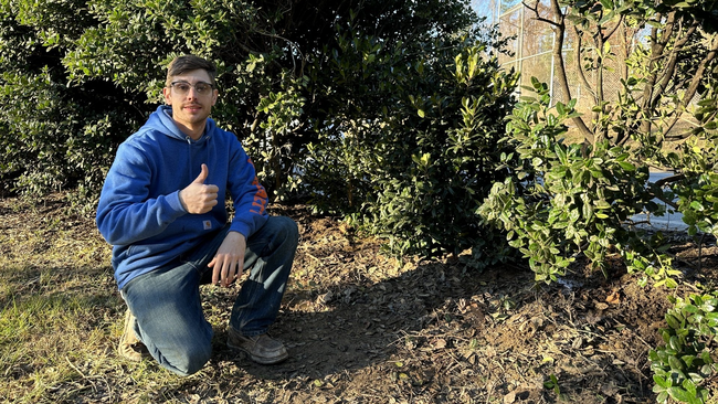 james thompson kneeling in front of hedge with thumbs up