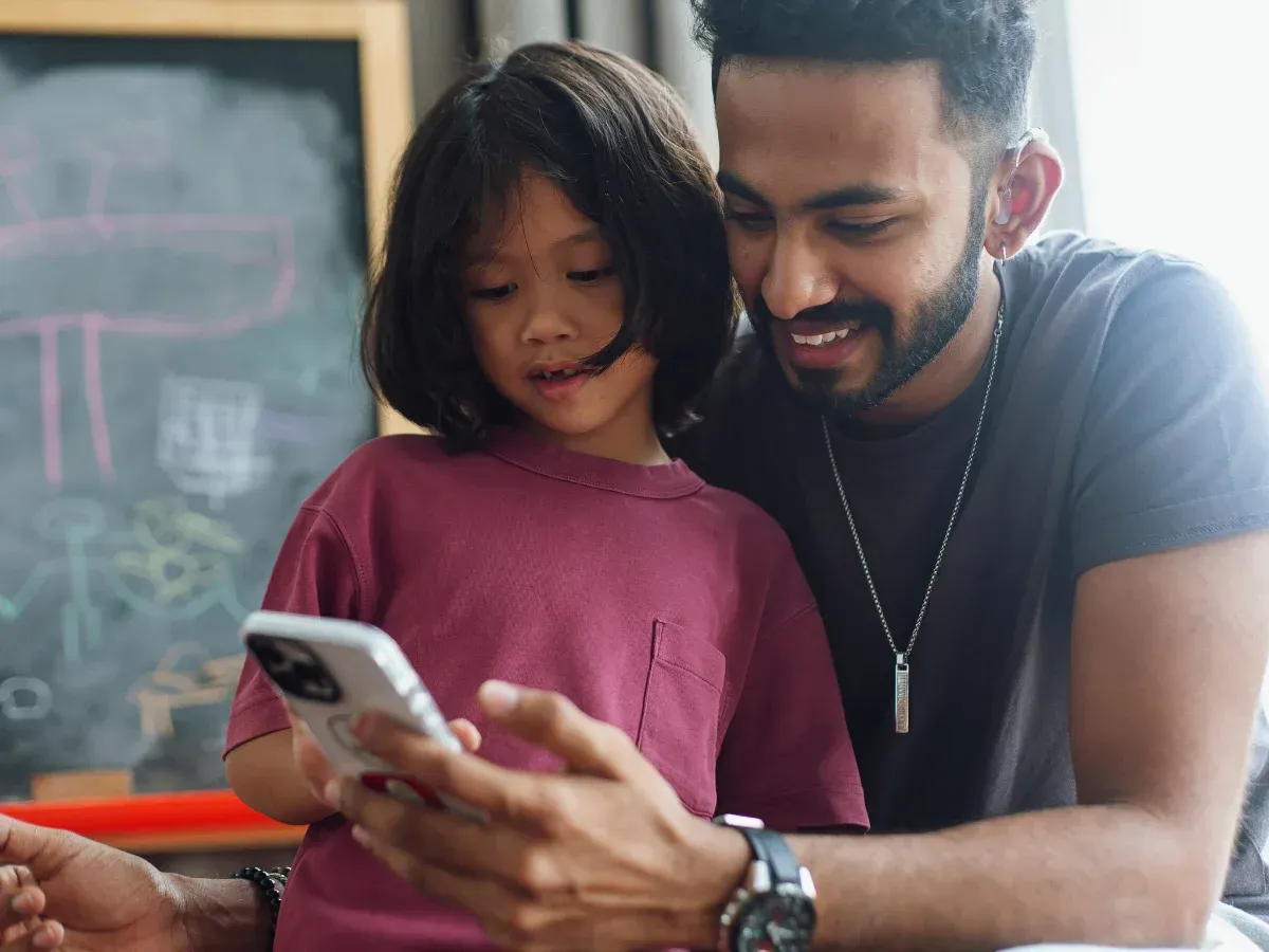 A man wearing a hearing aid watches a video on his phone with his daughter.