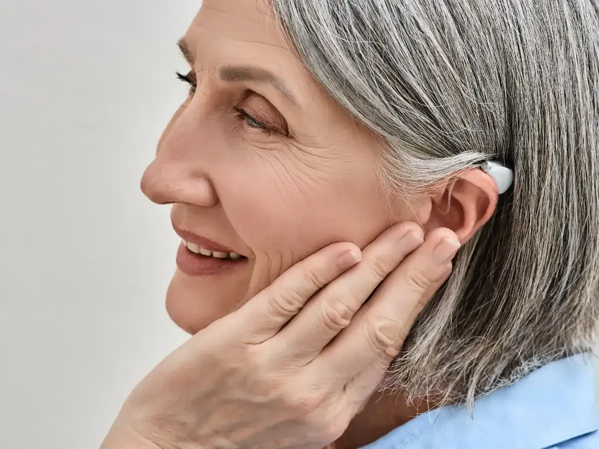 An elderly woman smiles while touching her hearing aid.
