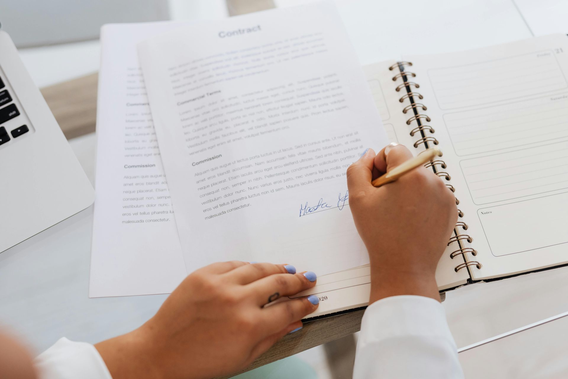 Person signing a contract with a pen on a desk. Notebook and laptop are present.