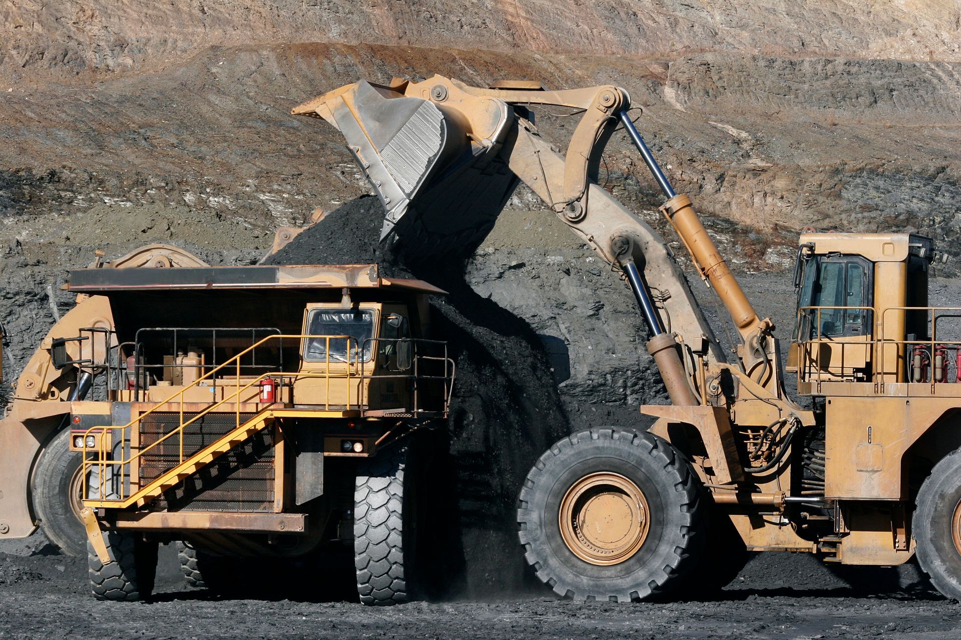 Excavator loads material into large mining truck at open-pit site with rocky terrain.
