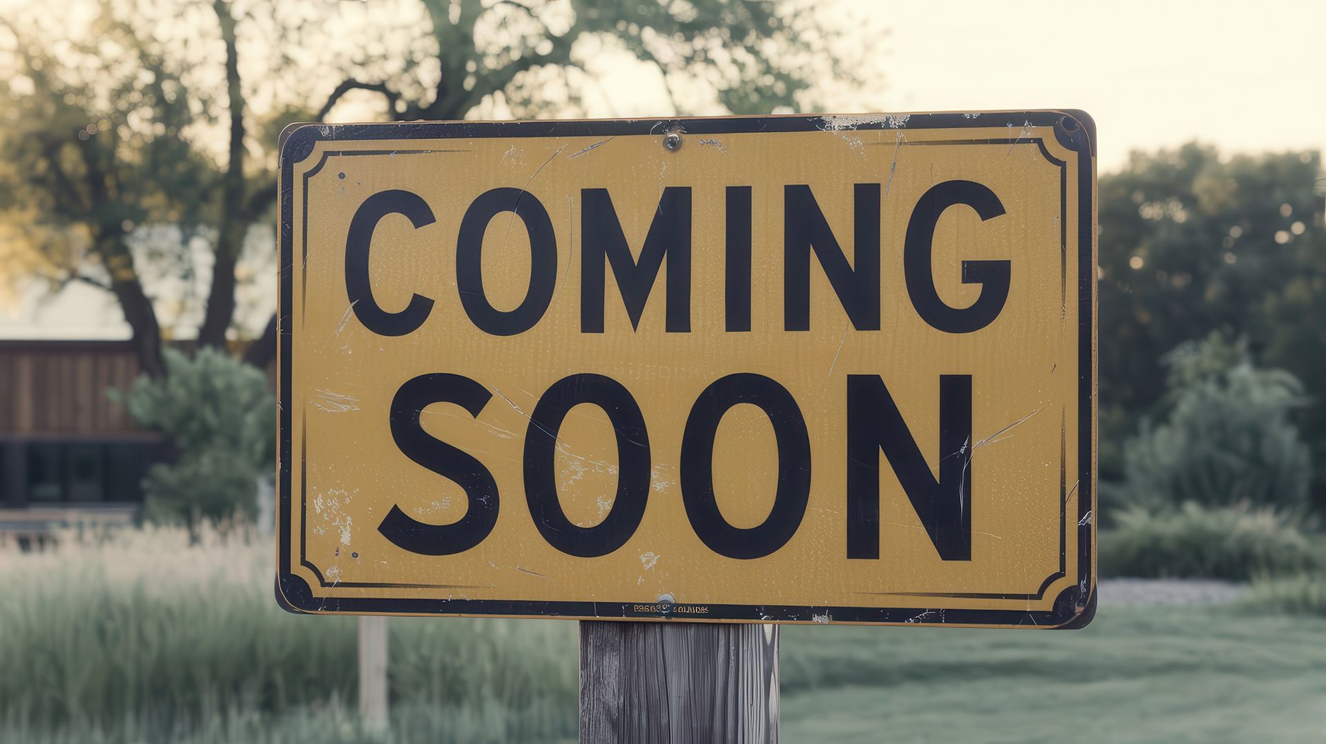 Weathered yellow sign reading “COMING SOON” on a post in a grassy outdoor area.