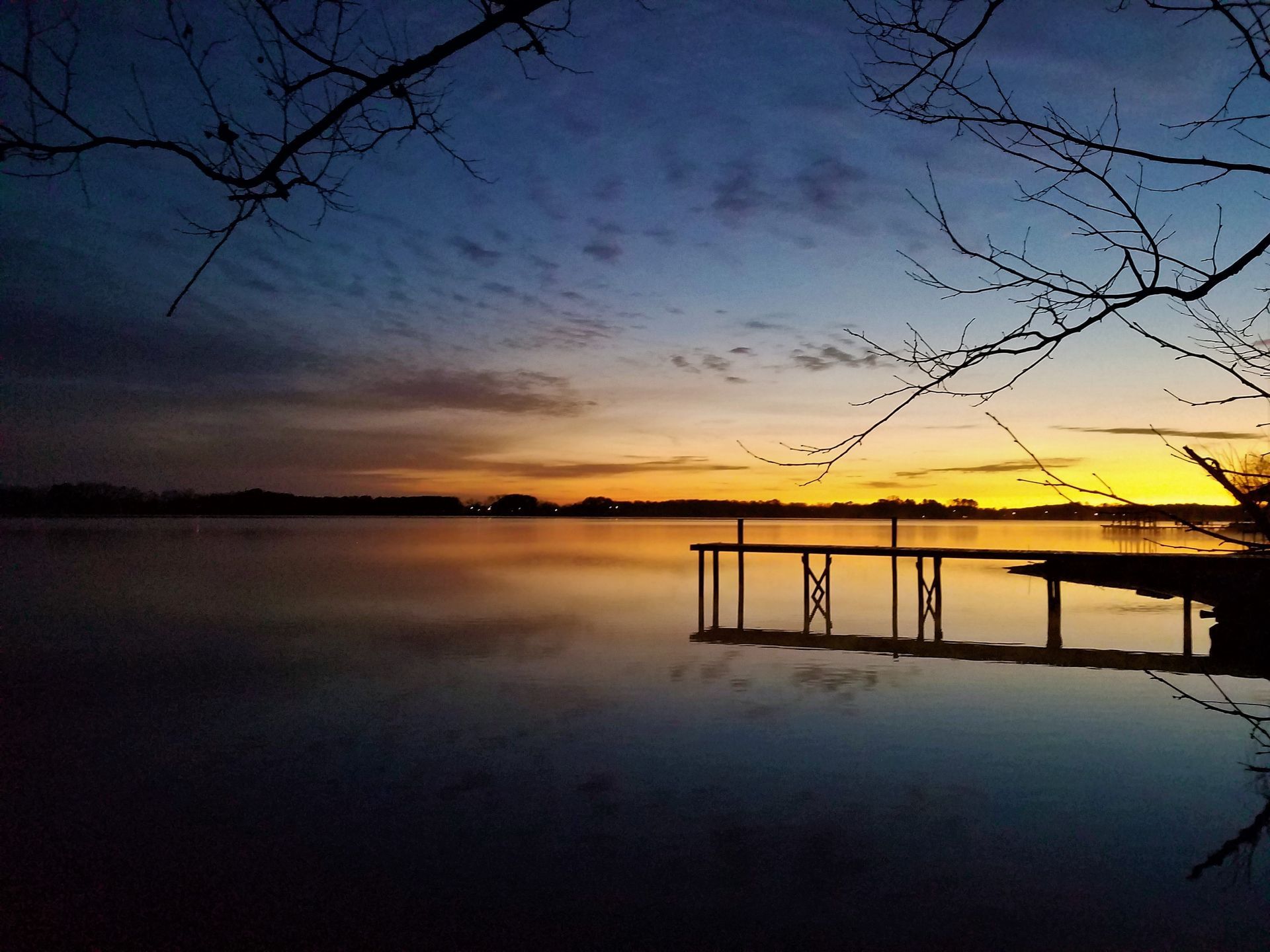 Sunset over calm lake with silhouetted dock and bare tree branches