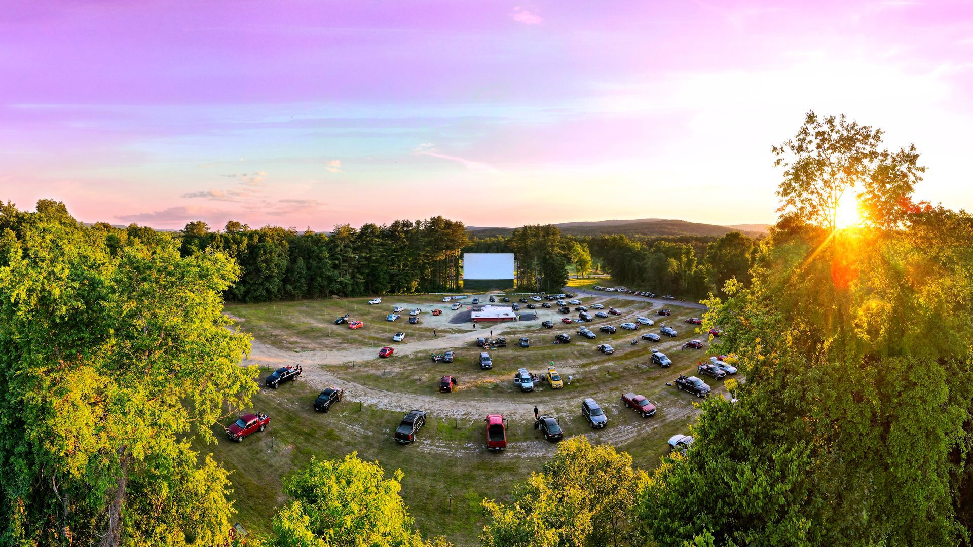 Outdoor drive-in movie theater at sunset, with cars parked facing a large screen under a pink sky