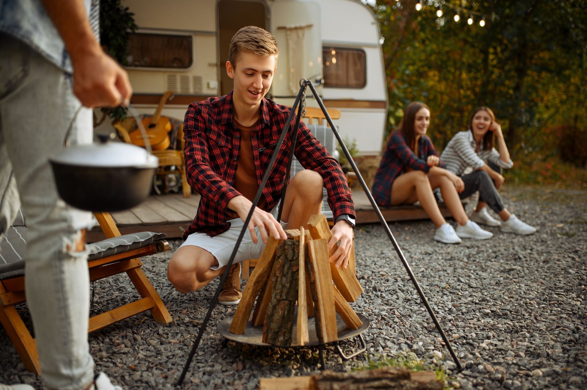 Friends roasting marshmallows around a campfire beside a camper at dusk