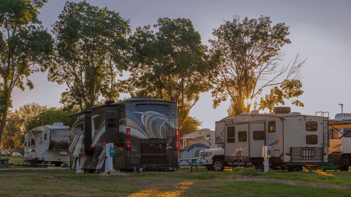 Camper trailers parked under trees at sunset, with a person standing beside a black RV.