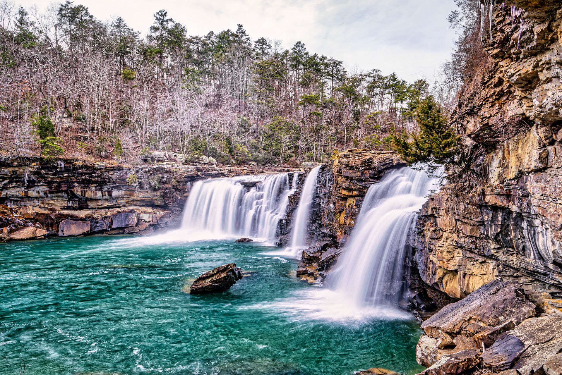 Twin waterfalls pouring into a turquoise pool beside rocky cliffs and trees