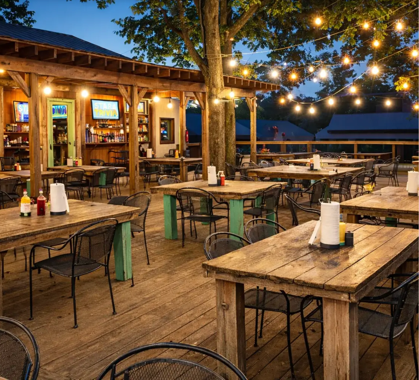 Outdoor wooden dining deck with empty tables, string lights, and a lit bar at dusk