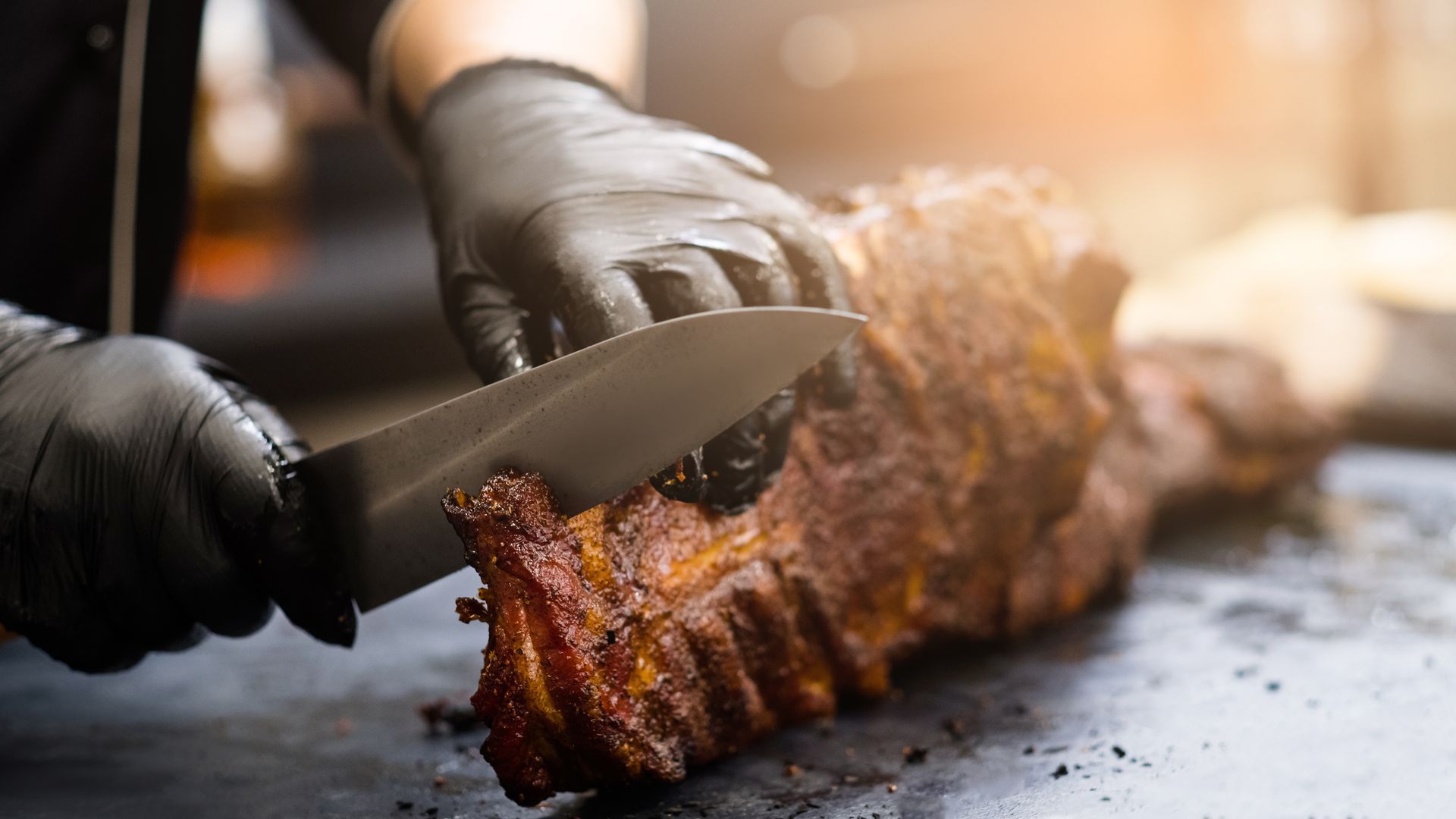 Gloved hands slicing a roasted rack of meat on a cutting board with a chef’s knife
