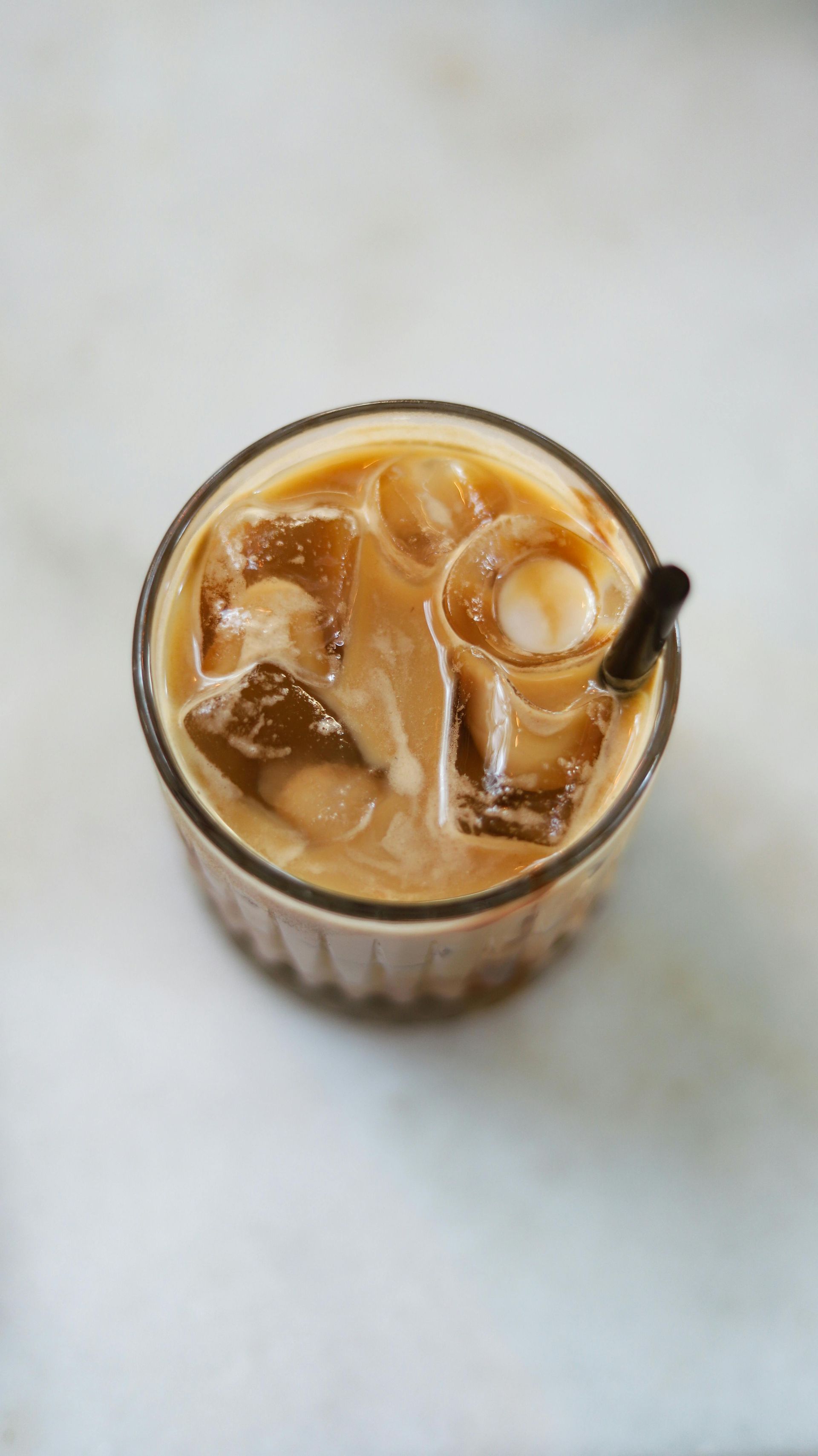 Top-down view of an iced coffee in a clear glass with a black straw on a light marble table