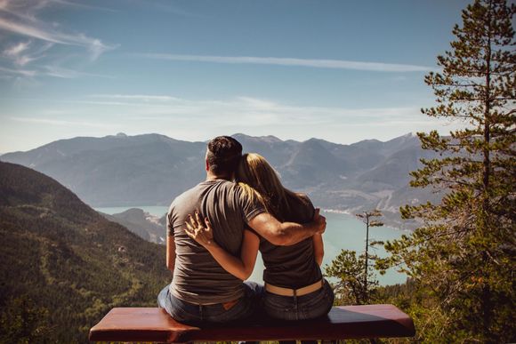 Two people sit on a bench overlooking a lake and mountains under a clear sky.