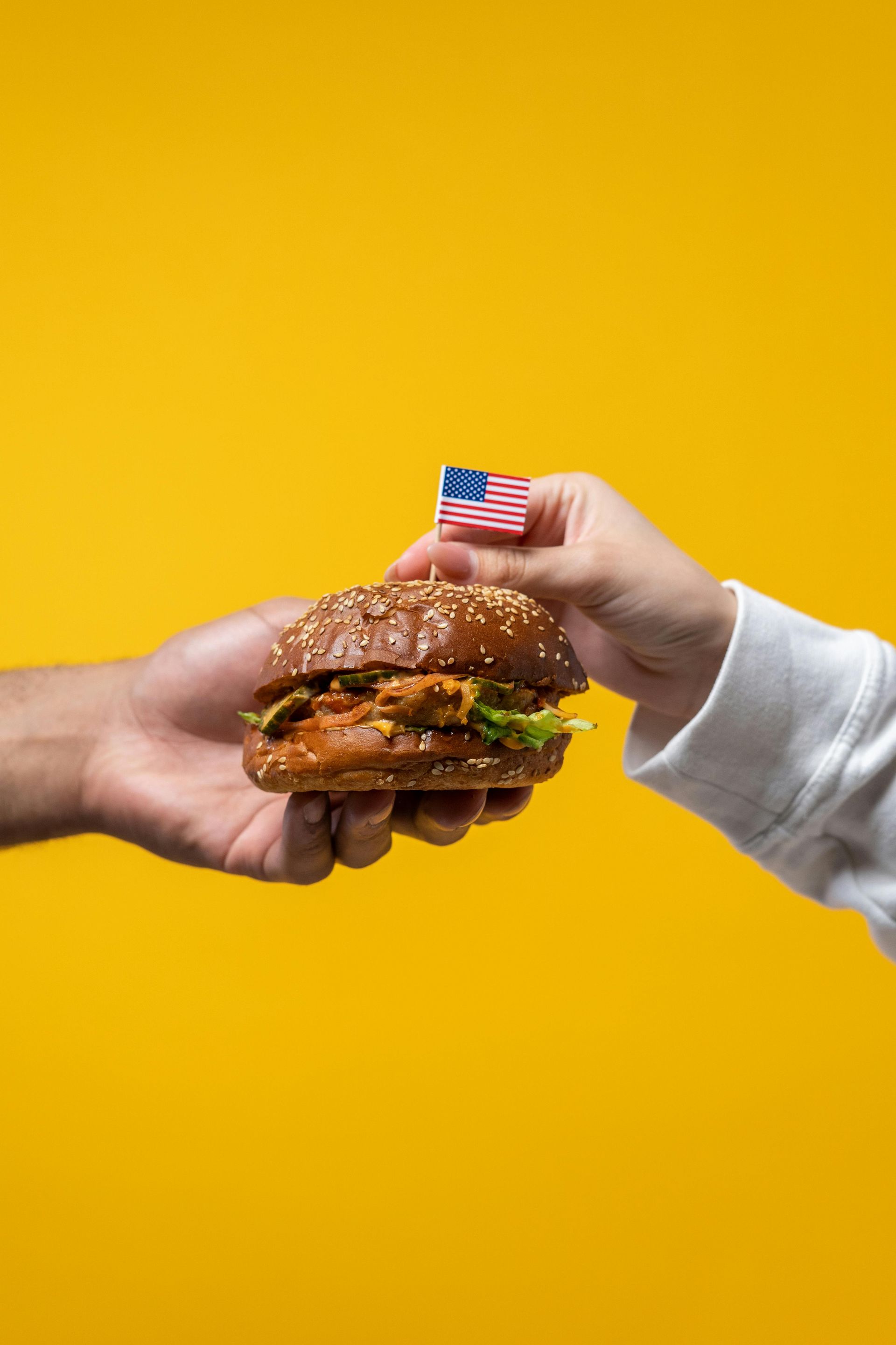 Two hands sharing a burger with a small American flag on a yellow background