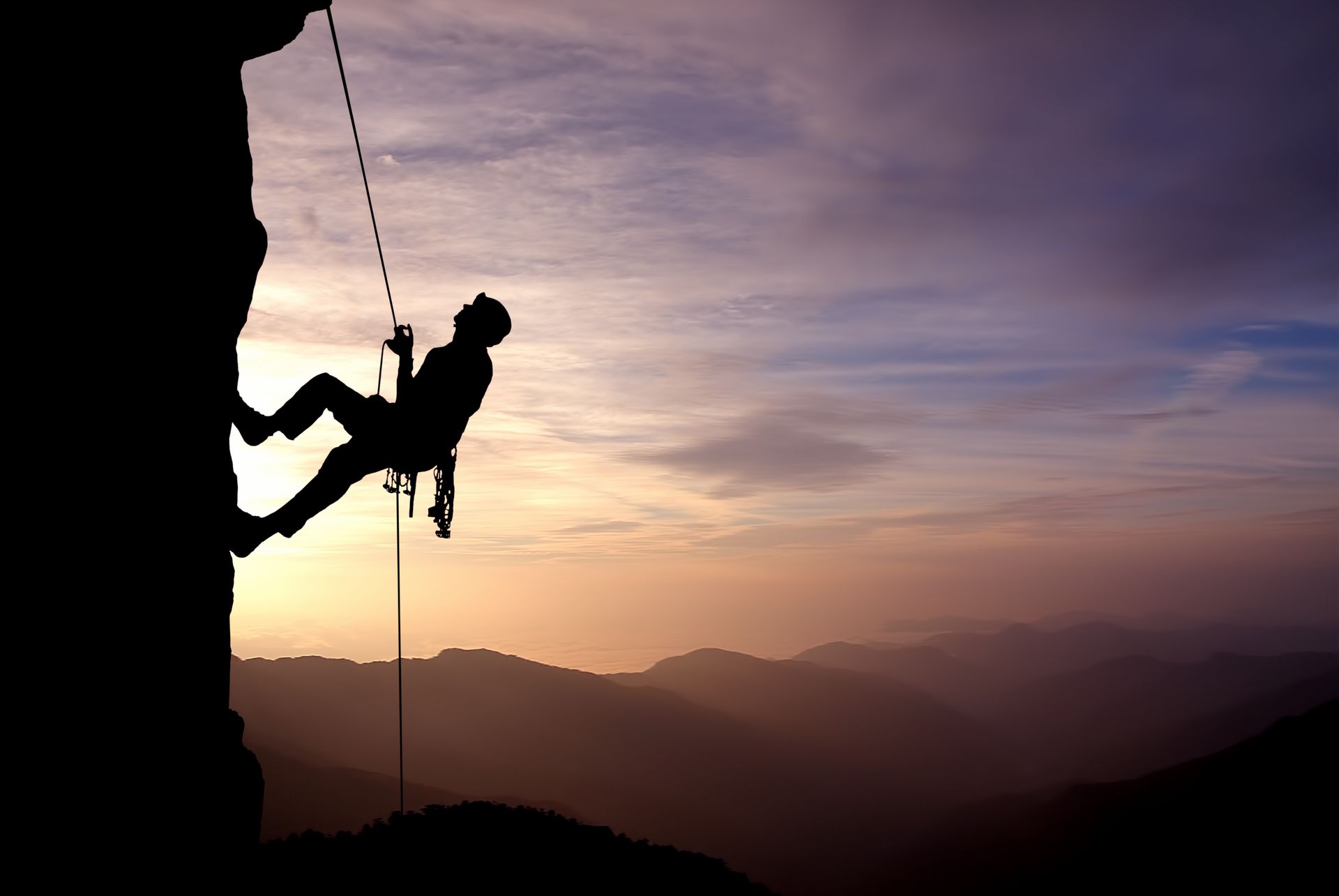 Silhouette of a rock climber descending a cliff at sunset, with mountains in the background