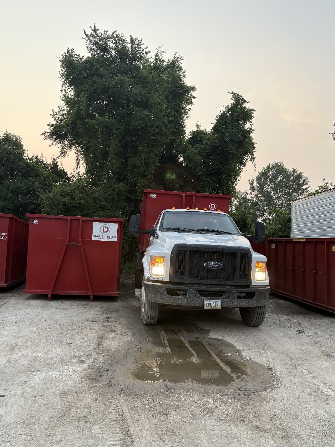 White Ford truck parked in front of red dumpsters; trees and sky in the background.