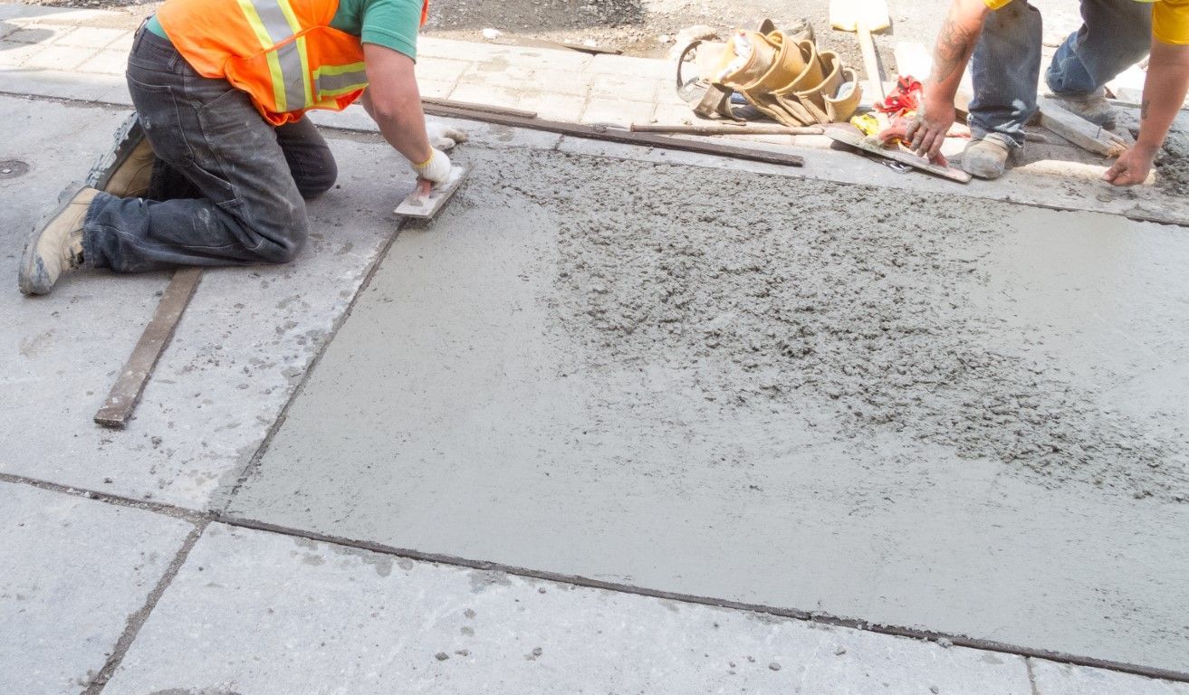 Two construction workers in safety vests smooth wet concrete on a sidewalk.