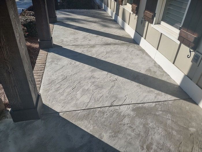 Sunlight casts geometric shadows from wooden posts onto a textured concrete walkway next to a building with a beige exterior.