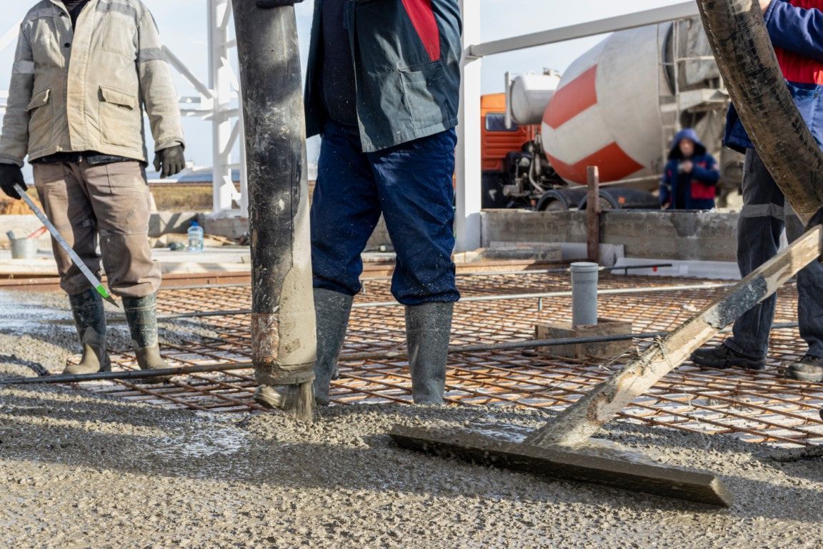 Construction workers in boots pour concrete onto metal rebar, smoothing it with a tool.