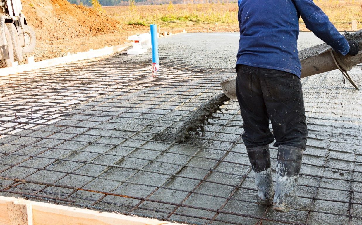 A worker in blue jacket and rubber boots pours concrete over a rebar grid for a building foundation.