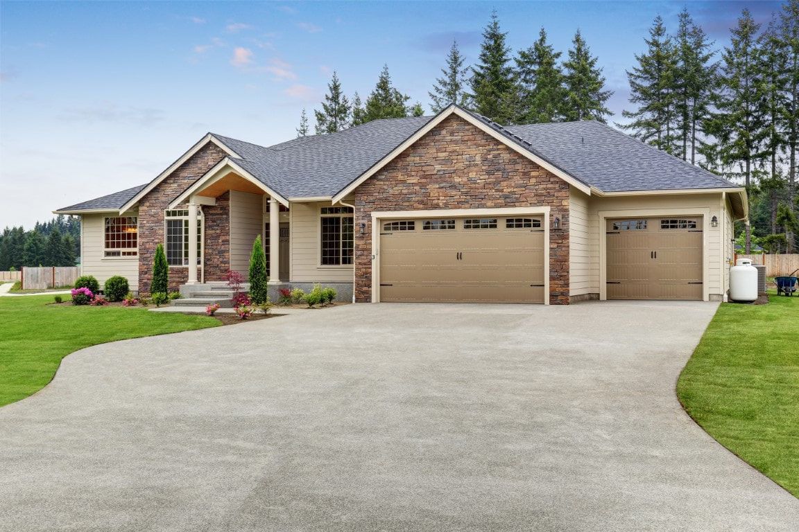 Single-story house with stone accents and large garage, surrounded by a manicured lawn and framed by tall evergreen trees.