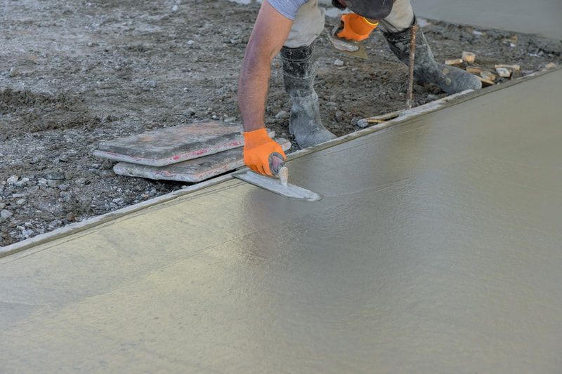 Worker in orange gloves smoothing wet concrete on a construction site. The surface is freshly poured.